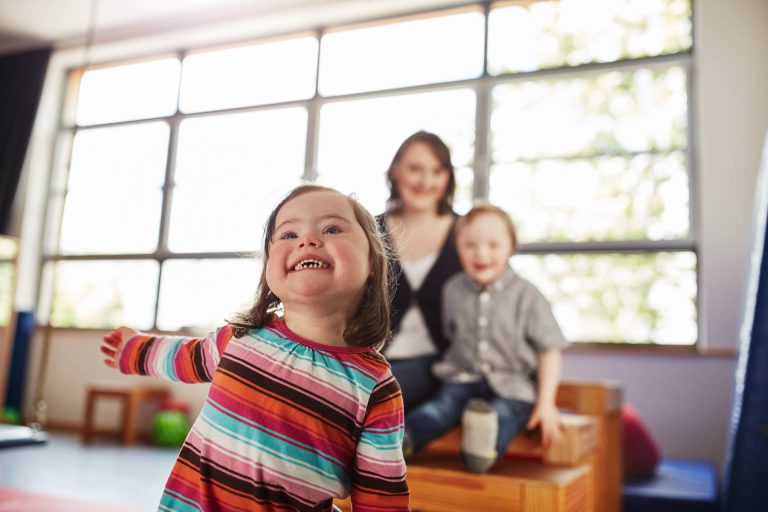 Zwei Kinder spielen mit ihrer Erzieherin. Das kleine Mädchen schaut den Betrachter direkt an.