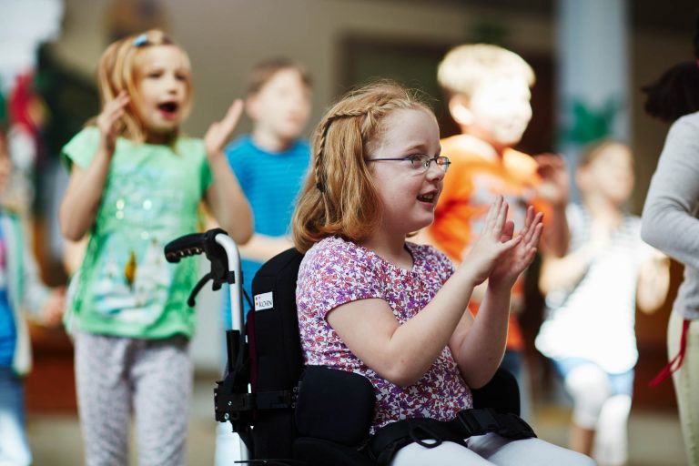 Ein kleines, rothaariges Mädchen mit schulterlangen Haaren und einer Brille sitzt im Rollstuhl. Sie sitzt im Fokus, verschiedene Kinder sind im Hintergrund unscharf erkennbar. Die Stimmung ist locker und gelöst, sie klatscht in die Hände.